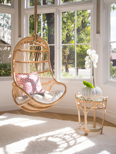 Kouboo Hanging Rattan Swing Chair With White Seat Cushion Hanging Next To The Window In Sunroom